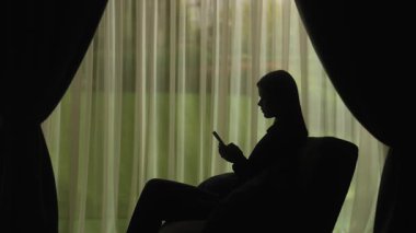 Kanepede oturmuş akıllı telefondan mesaj yazan bir kadının silueti. Perdeleri olan yumuşak ışıklı bir pencerenin önündeki kadın. Ruh sağlığı konsepti.