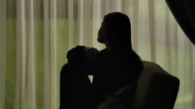 Yumuşak ışıklı perdelerin önünde oturan bir kadının silueti üzgün, stresli ve içe dönük ifade ediyor. Akıl Sağlığı Konsepti.