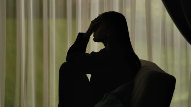Kanepede oturan ve ellerini yüzünü örten bir kadının silüeti. Perdeleri olan, pencerenin önünde sıkıntılı bir kadın, stres ve depresyonu ifade ediyor. Akıl Sağlığı Konsepti.