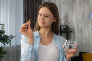 Bir kadın, sıcak bir yuva ortamında elinde bir bardak su tutarken şüpheli bir bakışla sarı bir kapsülü yakından inceliyor. Sağlıklı yaşam konsepti, cilt ve saç için vitamin almak..