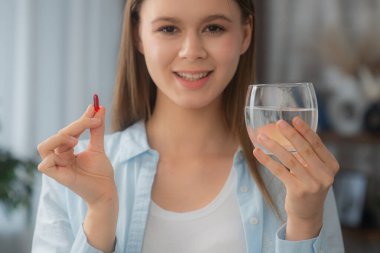 Bir elinde kırmızı bir hap, diğer elinde bir bardak su tutarken gülümseyen genç bir kadın, sağlık ve sağlığı temsil ediyor. Sağlıklı yaşam konsepti, cilt ve saç için vitamin almak..