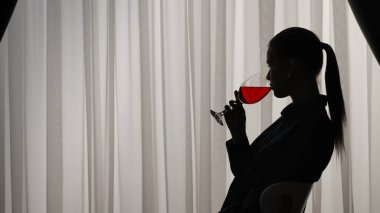 Kanepede oturan ve bardaktan kırmızı şarap içen bir kadının silueti. Yumuşak ışıklı pencerenin önünde perdeli bir kadın. Ruh sağlığı konsepti.