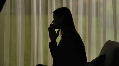 Kadın silueti, bir kanepede oturuyor ve önünde avuç içleri katlanmış dua ediyor. Yumuşak ışıklı pencerenin önünde perdeli bir kadın. Ruh sağlığı konsepti.