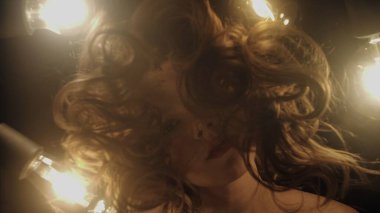 Dönen saçlar ve parıldayan ampullerle çevrili bir kadının gerçek üstü yakın çekimi gizemli ve sanatsal bir atmosfer yaratıyor..