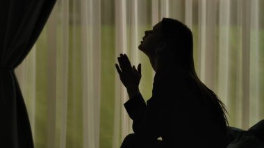 Kadın silueti, bir kanepede oturuyor ve önünde avuç içleri katlanmış dua ediyor. Yumuşak ışıklı pencerenin önünde perdeli bir kadın. Ruh sağlığı konsepti.