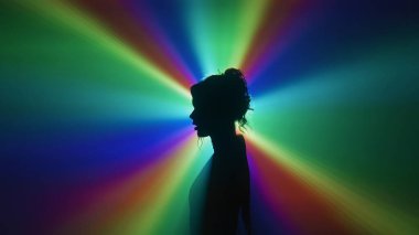 Bir kadının dramatik bir silueti canlı bir gökkuşağı ışık projeksiyonuna karşı duruyor, gizemli, sanatsal bir kompozisyonda vücudunun ana hatlarını belirten parlak bir renk patlaması yaratıyor. Yan görünüm.