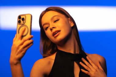 Genç bir kadın akıllı telefonuyla selfie çekerken yakalandı. Parlak mavi arka plan, onun parıldayan ifadesini vurgulayarak, sahneye modern ve canlı bir dokunuş ekler..