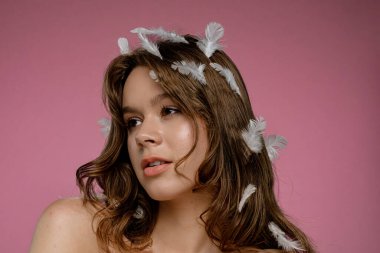 Pembe arka plan stüdyo ortamında saçında beyaz tüylerle poz veren doğal güzelliğe sahip bir kadın model. Şefkat, saflık ve güzellik kavramı..