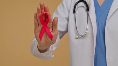 Tarafsız bej arka planda steteskoplu beyaz önlüklü doktor, farkındalığın sembolü olarak pembe kurdeleyle elini gösteriyor. Göğüs kanseri farkındalık günü, kadın sağlığı ve toplum kavramı.