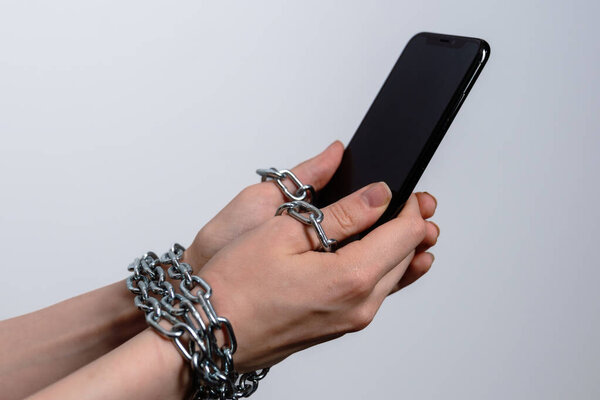 Женская рука, завернутая в металлические цепи держит смартфон. Концепция одержимости и зависимости от цифровых гаджетов и социальных сетей.