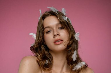 Pembe arka plan stüdyo ortamında saçında beyaz tüylerle poz veren doğal güzelliğe sahip bir kadın model. Şefkat, saflık ve güzellik kavramı..