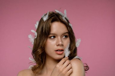 Bir kadın stüdyo ortamında yüzünün yanında beyaz bir tüyle poz veriyor. Düşen beyaz tüylü pembe arka plan. Çekim sakin ve yumuşak bir atmosfer sağlıyor..