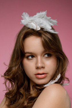 Doğal güzelliğe sahip genç bir kadın model pembe bir arka planda kafasında tüylerle poz veriyor. Şefkat, saflık ve güzellik kavramı..