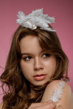 Doğal güzelliğe sahip genç bir kadın model pembe bir arka planda kafasında tüylerle poz veriyor. Şefkat, saflık ve güzellik kavramı..