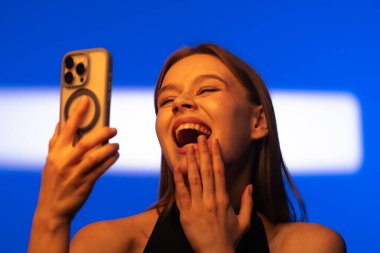 Genç bir kadın, parlak mavi bir arka planda akıllı telefonuyla neşeyle selfie çekiyor..