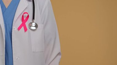 Beyaz önlüklü, steteskoplu ve pembe kurdeleli bir doktor tarafsız bir arkaplanı işaret ediyor. Göğüs kanseri farkındalık günü konsepti.