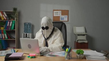 İşte hayalet gibi giyinmiş biri. Beyaz çarşaflı cadılar bayramı işadamı hayaleti büro masasında oturur dizüstü bilgisayarda çalışır, mutlu ifade kazanan hareketi gösterir. Cadılar Bayramı iş konseptiniz kutlu olsun..