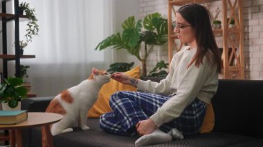 Kanepede bacak bacak üstüne atmış genç bir kadın turuncu ve beyaz kedisine kıyak yapıyor. Sahne sakin, rahat ve doğal ışık ve yeşillikle dolu..