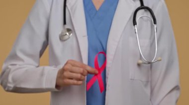 Bej arka planda beyaz önlüklü bir doktor, farkındalığın sembolü olarak kameraya pembe kurdeleli bir el uzatıyor. Göğüs kanseri farkındalık günü, kadın sağlığı ve toplum konsepti.