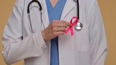 Steteskop ve pembe kurdeleli beyaz önlüklü doktor, farkındalığın sembolü olarak kurdeleyi işaret ediyor. Göğüs kanseri farkındalık günü konsepti.