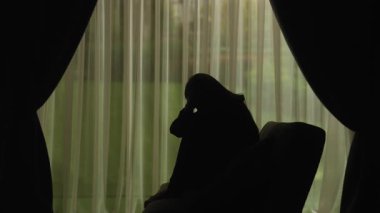 Yumuşak ışıklı perdelerin önünde oturan bir kadının silueti üzgün, stresli ve içe dönük ifade ediyor. Akıl Sağlığı Konsepti.