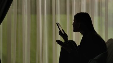 Kanepede oturmuş akıllı telefonda agresif bir şekilde konuşan bir kadının silueti. Perdeleri olan bir pencerenin önündeki kadın. Ruh sağlığı kavramı.