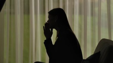 Kanepede oturan ve birinden bir şey isteyen bir kadının silueti. Yalvarma jesti ile ellerini kavuşturuyor. Yumuşak ışıklı pencerenin önünde perdeleri olan bir kadın. Akıl sağlığı kavramı.