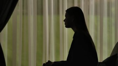 Kanepede oturan ve biriyle iletişim kuran bir kadının silueti. Perdeleri olan pencerenin önündeki kadın. Akıl sağlığı kavramı.