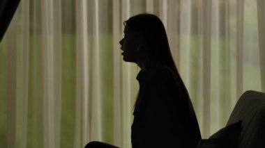 Yumuşak ışıklı perdelerin önünde oturan bir kadının silueti üzgün, stresli ve içe dönük ifade ediyor. Akıl Sağlığı Konsepti.