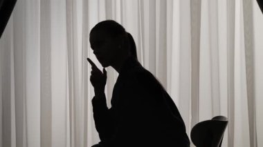 Yumuşak ışıklı perdelerin önünde oturan bir kadının silueti. Kadın bir şey düşünüyor, endişeleniyor ve soruna bir çözüm bulamıyor..