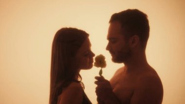 Hassas bir anı paylaşan bir çiftin silüetli aşk hikayesi aralarındaki yakınlığı ve sıcaklığı vurgular. Bir erkek bir kadına çiçek verir..