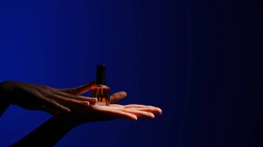 Bir el nazikçe sunar yumuşak ışığın altında bir oje şişesi koyu mavi bir arka plana karşı, zarafeti vurguluyor..