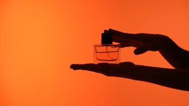 Ellerin silueti zarif bir şekilde bir şişe parfümü çarpıcı bir turuncu zemine tutuyor..