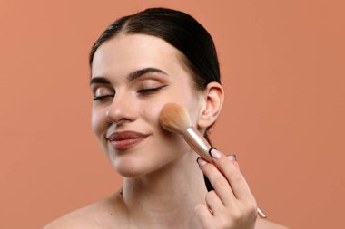 Yanağında makyaj fırçası ile gülümseyen bir kadın yumuşak turuncu arka planda doğal bir görünüm sergiliyor..
