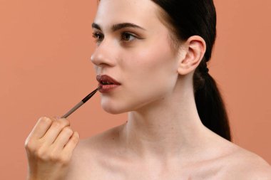 Şeftali arka planına makyaj fırçası sürerek ruj süren bir kadın. Odak noktası hassasiyet ve dudak rengi uygulaması.