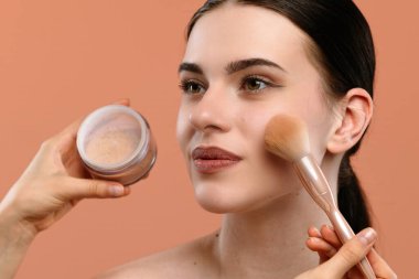 Bir kadın yumuşak bir fırçayla gevşek yüz pudrası sürer. Arka plan şeftali renginde..