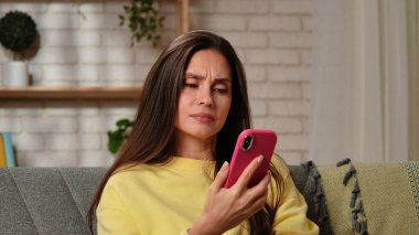 Kadın evde boş vakit geçiriyor. Genç bir kadın, oturma odasında kanepede oturmuş elinde akıllı bir telefonla sosyal medyayı karıştırıyor ve kafası karışık bir ifade takınıyor..