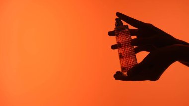 Ellerin silueti zarif bir şekilde bir şişe parfümü çarpıcı bir turuncu zemine tutuyor..