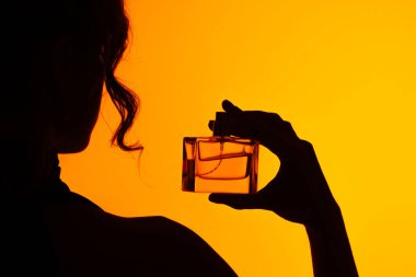 Bir kadının silüeti bir parfüm şişesini canlı turuncu arka planda tutuyor, zarafet ve gizemi çağrıştırıyor..