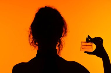 Parfüm şişesi tutan bir kadının silüeti canlı turuncu bir arkaplanda, zarafeti ön plana çıkarıyor..