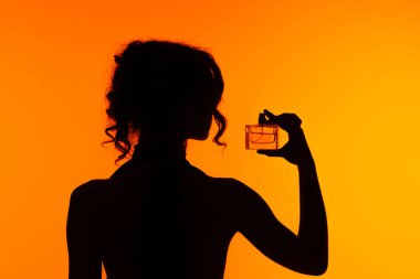 Parfüm şişesini parlak turuncu arka planda tutan bir kadının silüeti zarafet ve gizemi çağrıştırıyor..