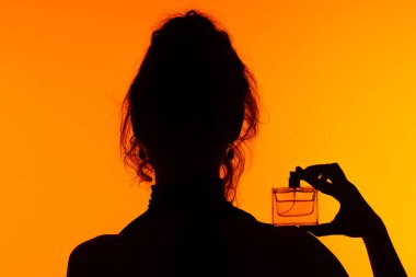 Zarif bir parfüm şişesi tutan bir kadının büyüleyici silüeti, portakal arkaplanına yaslanmış, zarafet ve gizemi çağrıştırıyor..