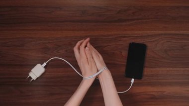 Telefon şarj kablosu ile bağlanan eller modern cihazlara ve teknolojik kısıtlamalara bağımlılığı simgeler.