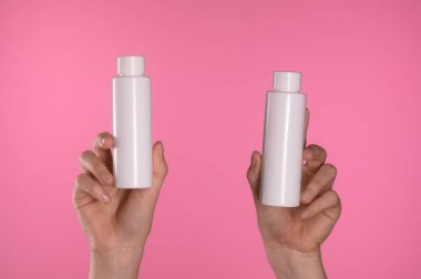 İki el aynı beyaz plastik şişeleri pembe bir zemin üzerinde tutuyor, ürün ambalaj simetrisini vurguluyor..