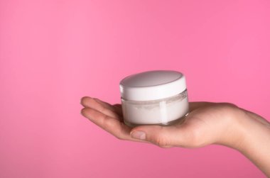 Canlı pembe bir arka planda beyaz krema kavanozunu tutan bir el. Cilt ve güzelliği sembolize ediyor..