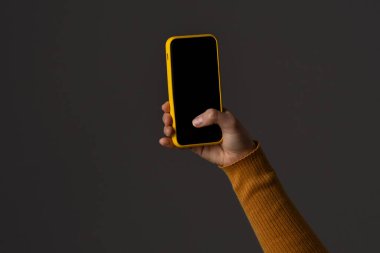 Canlı sarı çantalı akıllı telefonu tutan bir el. Arka plan koyu bir gölgedir, telefon rengini arttırır..