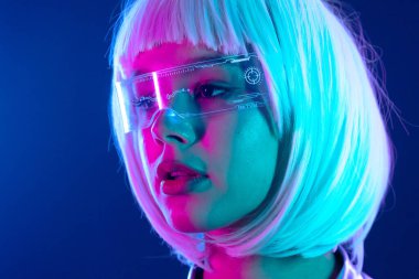 Fütürist bir kadının portresi, parlak neon ışıkların altında holografik akıllı gözlük takıyor. Mavi ve pembe ışıklar beyaz saçlarını ve metalik kıyafetlerini ön plana çıkarıyor. Siber punk estetiğini çağrıştırıyor..