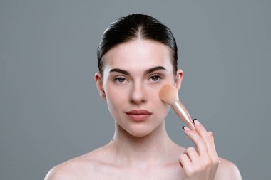 Genç bir kadın, yanak bölgesine odaklanmak için fırça kullanarak makyaj yapıyor. Minimalist arka plan tekniğini ve doğal özelliklerinin güzelliğini vurguluyor..