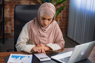 Orta Doğulu bej tesettürlü Müslüman kadın çatı katında masa başında çalışıyor. Kadın hesap makinesindeki verileri hesaplıyor ve masadaki çizelgelerle karşılaştırıyor..