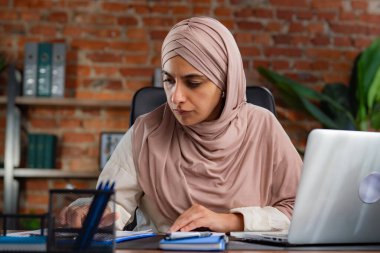 Orta Doğulu bej tesettürlü Müslüman kadın çatı katında masa başında çalışıyor. Kadın hesap makinesindeki verileri hesaplıyor ve masadaki çizelgelerle karşılaştırıyor..